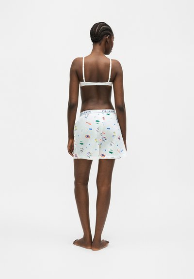 Γυναίκα με πλεξούδες, όρθια ξυπόλητη με την πλάτη, φοράει λευκό σουτιέν και λευκά σορτς Polo Ralph Lauren με πολύχρωμα σχέδια.