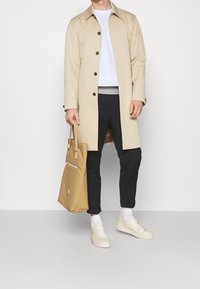 Gabardina beige con cierre de botones, suéter blanco, pantalones azul marino, zapatillas crema y un bolso tote marrón claro con detalle de cremallera.