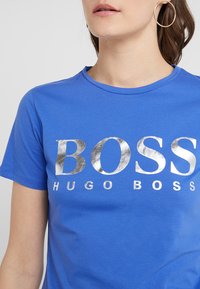 Blå bomullströja med rund halsringning och silvermetallisk text "BOSS" med "HUGO BOSS" under, slät textur.