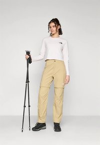 Beige outdoor pants with a zippered pocket, paired with a light long-sleeve shirt. Holding black trekking poles, wearing black hiking shoes.