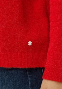 Czerwony sweter z miękkiej, teksturowanej tkaniny, z prążkowanym wykończeniem i małym białym guzikiem jako akcentem. Noszony na ciemnoniebieskich dżinsach.