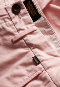 Pantalones cortos de algodón rosa con un acabado texturizado, que presentan un botón negro y una pequeña etiqueta negra con el nombre de la marca en la cinturilla.