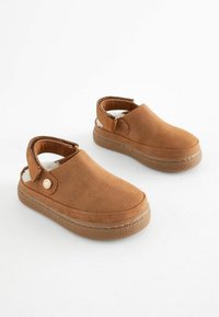 Chaussures brunes à enfiler en matériau doux, avec une semelle texturée, un talon ouvert et un petit emblème sur le côté.