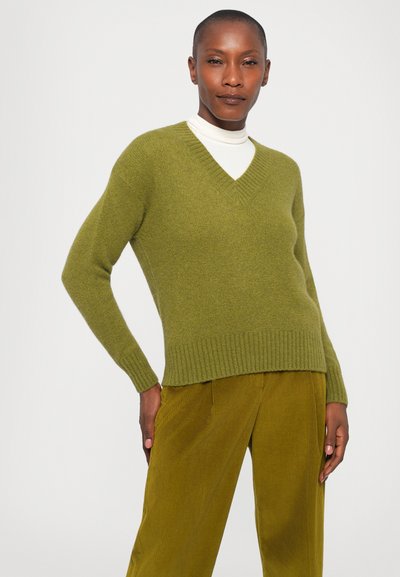 Green knit sweater with a V-neck, ribbed cuffs, and hem. Worn over a cream turtleneck, paired with textured olive green corduroy pants.