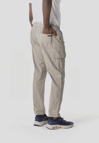 Hombre con pantalones cargo beige y camisa blanca, de pie con la mano derecha en el bolsillo trasero, vistiendo zapatillas deportivas azul marino y beige.