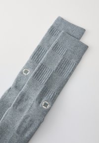 Calze grigie in maglia con texture a coste e un motivo geometrico intrecciato, dotate di una patch rotonda con logo sul lato.