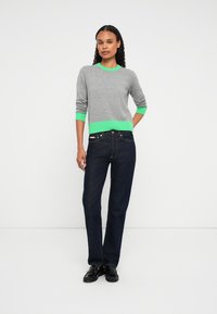 Mulher em pé vestindo um suéter cinza com detalhes em verde brilhante, jeans reto azul-escuro e mocassins pretos, contra um fundo branco liso.