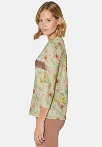 Langärmliges Oberteil mit floralem Muster in Grün, das Schattierungen von rosa und gelben Blumen zeigt. Brauner Akzentstreifen mit dem Text "SPORTS."