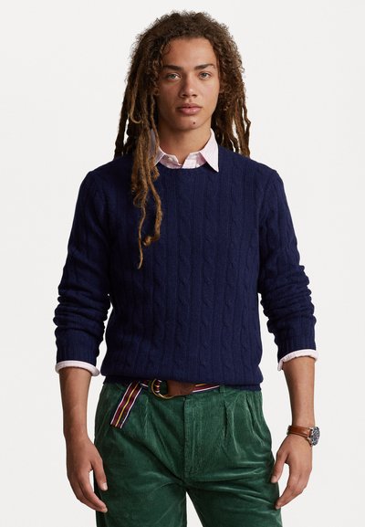 Pull en maille torsadée marine à manches longues, porté par-dessus une chemise rose clair à col. Associé à un pantalon en velours côtelé vert et une ceinture multicolore.