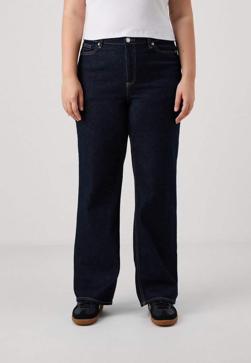 Pantalones vaqueros de pierna ancha azul oscuro con cintura alta, que cuentan con cinco bolsillos y costuras visibles. Combinados con zapatillas negras sobre una superficie lisa.