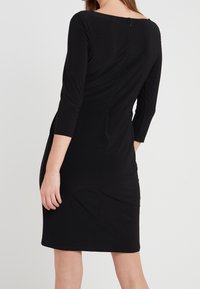 Kobieta w dopasowanej, czarnej sukience do kolan z rękawami o długości trzech kwadransów, pokazana od tyłu na jednolitym jasnym tle.