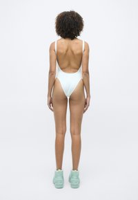 Femme debout de dos, portant un maillot de bain une pièce blanc et des bottes cheville vert menthe sur un fond clair et uni.