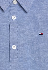 Jasnoniebieska koszula z guzikami, wykonana z teksturowanej tkaniny, z ostrym kołnierzykiem i białymi guzikami. Mały haftowany logotyp w kolorach czerwonym, białym i niebieskim.