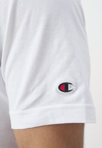Vit bomullströja med en svart, röd och vit broderad logotyp på vänster ärm. Kort ärm med en mjuk yta.
