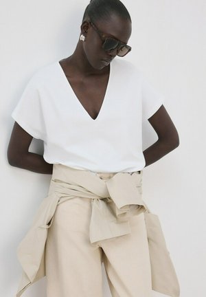 Frau trägt dunkle Sonnenbrille, weißes V-Ausschnitt-Shirt, beige Hose mit einer passenden beigen Jacke, die um die Taille gebunden ist, vor einfarbigem Hintergrund.