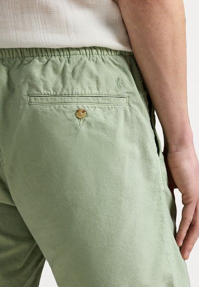 Un pantalon en coton vert clair avec une taille élastique et une poche arrière avec un bouton en bois. Un branding subtil est visible sur le tissu.