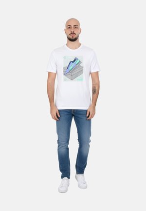 Uomo con barba che indossa una maglietta bianca con una grafica colorata di sneaker, jeans blu e scarpe da ginnastica bianche, in piedi davanti a uno sfondo semplice.