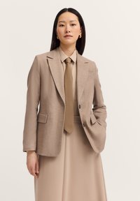 Hellbrauner eleganter Blazer mit Revers, zwei Vorderfächern und einem einzigen Knopfverschluss, getragen über einem beigen Hemd und einer Krawatte.