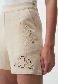 Beige Leinenkurze Hosen mit floralen Stickereien und Ausschnitten an den Seiten. Sie besitzen eine schmale Taille und einen sauberen Saum.