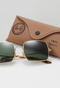 Occhiali da sole quadrati con montatura dorata e lenti verdi. Accompagnati da una custodia in pelle marrone texture con logo e chiusura a scatto.