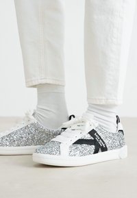 Hvite bukser og sokker med glitrende sølv sneakers med svarte sidestiper og hvite skolisser på et lyst gulv.
