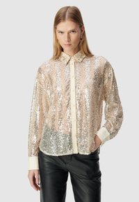 Donna con capelli biondi lisci che indossa una camicetta trasparente a maniche lunghe con paillettes, polsini e colletto color crema, abbinata a pantaloni neri in pelle.