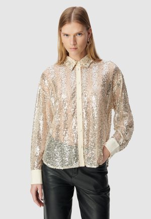 Woman with straight blonde hair wears a sheer, sequined long-sleeve blouse with cream cuffs and collar, paired with black leather pants.