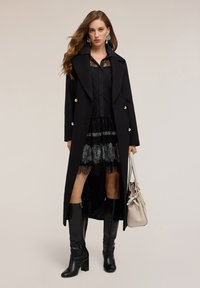 Lungo cappotto nero con bottoni dorati su un vestito di pizzo nero. Abbinato a stivali neri al ginocchio e a una borsa beige chiaro.