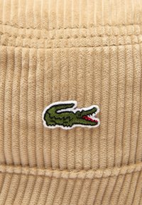Cappello bucket in corduroy beige con texture a righe verticali, arricchito da un logo di coccodrillo verde ricamato con dettagli rossi.