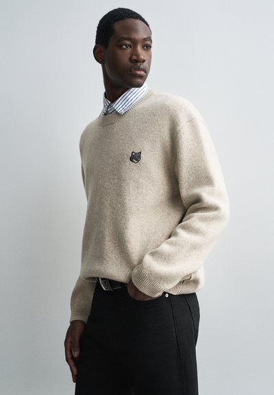 Beige gebreide trui met een ronde hals, voorzien van een klein zwart vossenembleem op de borst. Gekleed met een blauw-witte gestreepte shirt.