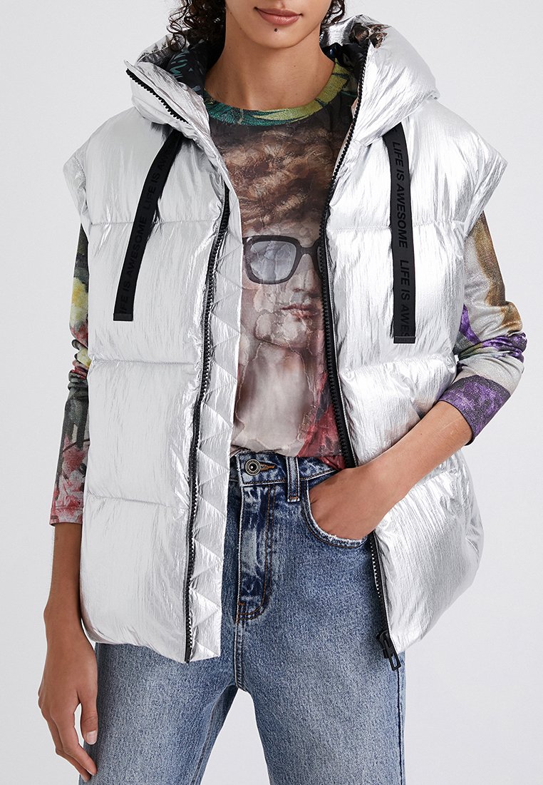Srebrna kamizelka puchowa o błyszczącej powierzchni, z zamkiem z przodu i czarnymi paskami. Noszona na kolorową koszulkę z długim rękawem i w połączeniu z dżinsami.