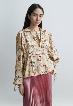 Ľahká béžová blúzka s kvetinovým vzorom, s viazaním na krku, dlhými rukávmi a voľným strihom, v kombinácii s plisovanou ružovou sukňou.