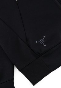 Chaqueta deportiva negra con una textura suave. Presenta un cuello con cremallera y puños en las muñecas, con detalles de marca en blanco cerca del dobladillo.