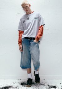 Grå grafisk t-shirt lagdelt over en orange ternet langærmet skjorte, parret med brede denimshorts og sorte sko med tykke såler.