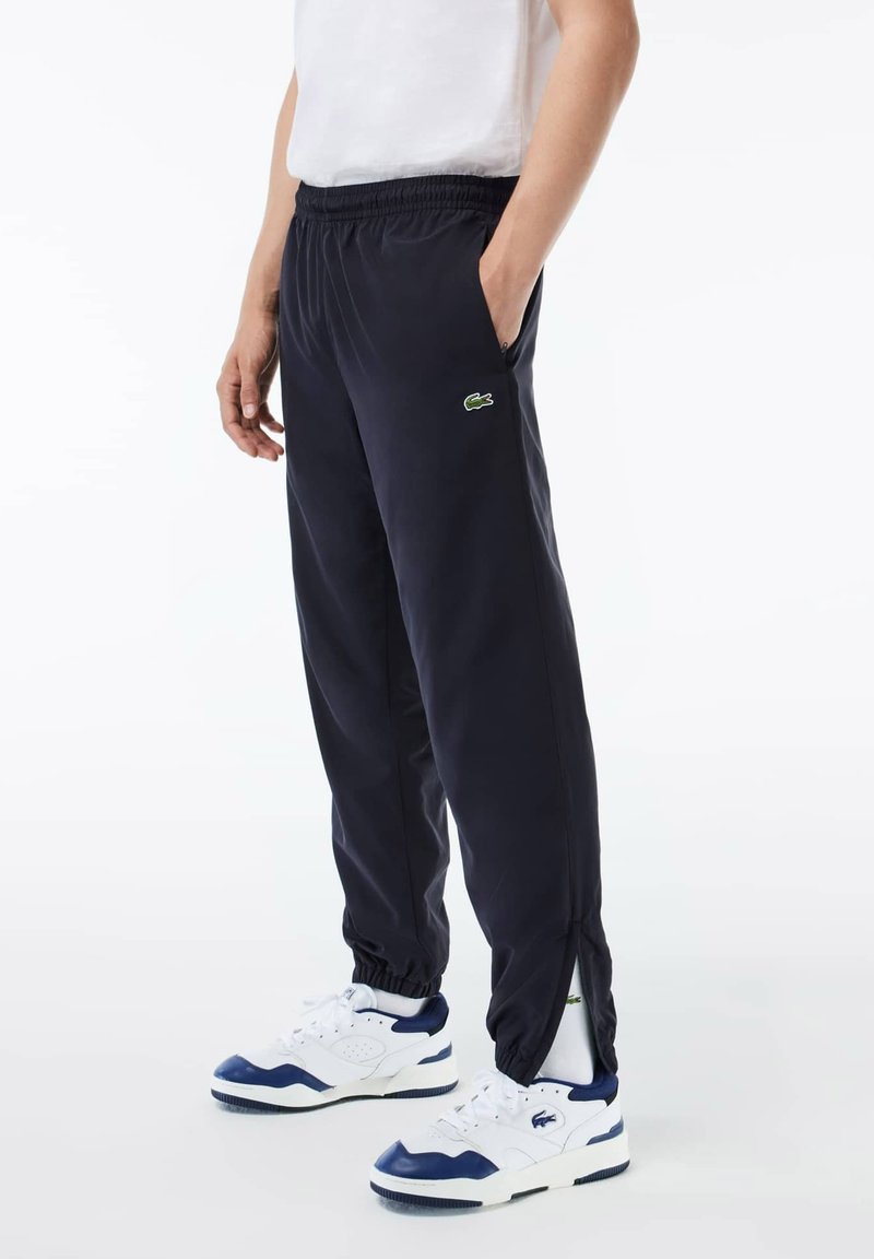 Pantalons de survêtement navy avec taille élastique, poches latérales et logo Lacoste en vert. Portés avec des baskets blanches avec des accents bleus.