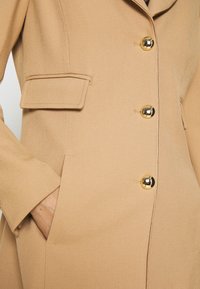 Cappotto tan con un design strutturato, caratterizzato da grandi bottoni dorati, una tasca sartoriale e una texture liscia.