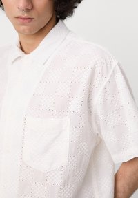 Weißes kurzärmliges Hemd mit Knopfleiste, das ein strukturiertes Design mit kleinen Ausschnitten, eine Tasche auf der linken Brust und einen spitzen Kragen aufweist.