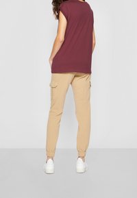 Vínová tričková košile s krátkým rukávem a volným střihem, doplněná bežovými cargo kalhotami s elastickými manžetami a bočními kapsami. Bílá sportovní obuv dotváří celkový vzhled.