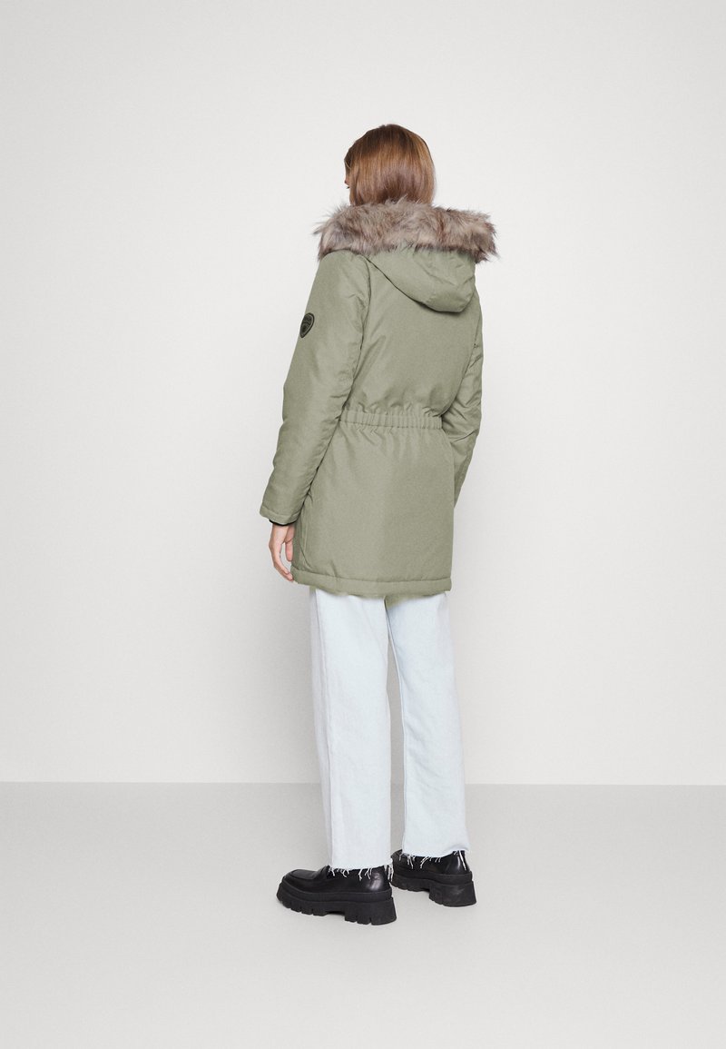 Parka matelassée vert olive avec capuche en fausse fourrure, taille cintrée et poches latérales, associée à un jean bleu clair et des bottes à plateforme noires.