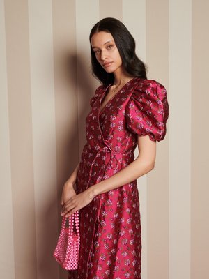Mujer con cabello largo y oscuro, vistiendo un vestido de manga abullonada rosa con flores y sosteniendo un bolso de cuentas rosa frente a un fondo rayado beige.