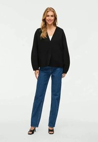 Melna lielizmēra kardigāna ar V veida kakla izgriezumu, saskaņota ar ziliem augsto jostasvidu taisnām biksēm un melniem sandalēm ar atvērtu purngalu. Gluda tekstūra.