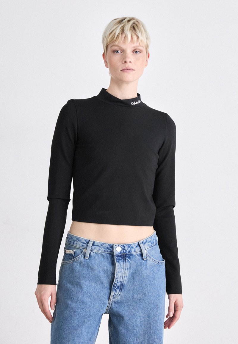 Crop top noir à manches longues avec un col montant et le logo Calvin Klein sur le col, associé à un jean en denim bleu clair taille haute.