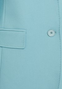 Il tessuto del blazer azzurro chiaro presenta un bottone singolo, dettagli cuciti e un design con tasca, con una texture liscia e una vestibilità sartoriale.