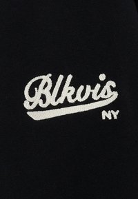 Czarna tkanina z białym haftowanym napisem "Blkvis NY" w kursywie z stylizowanym podkreśleniem. Haft pokazuje drobne szczegóły.
