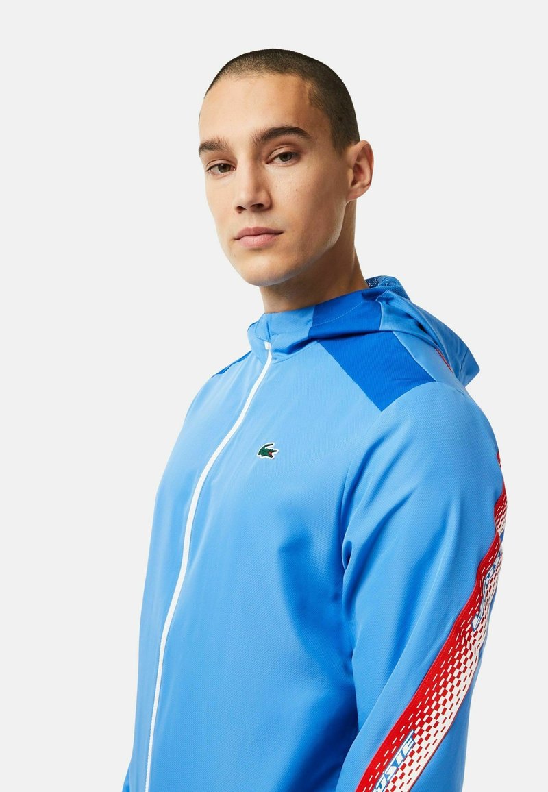 Veste à zip bleu clair avec capuche, présentant une finition texturée, des panneaux d'épaule bleus contrastants et un accent rouge et blanc à motifs sur la manche.