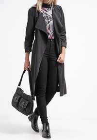 Long manteau noir à l'avant ouvert, porté sur un haut à motifs et un jean skinny noir. Bottines noires et sac à main noir avec des accents.