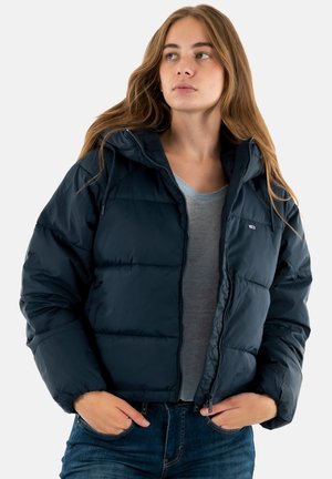 Junge Frau mit langen Haaren, die eine schwarze Steppjacke über einem grauen Shirt und blauen Jeans trägt, mit den Händen in den Taschen, die zur Seite schaut.