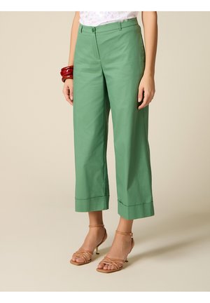 Donna che indossa pantaloni verdi a vita alta, larghi e corti, abbinati a sandali con tacco color cuoio con cinturini e a un braccialetto rigido rosso.