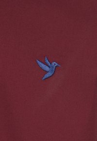 Bordeaux stof med en lille blå broderet kolibri med udstrakte vinger, centreret på materialet.