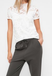 Blusa bianca in pizzo con maniche corte, motivi floreali, scollatura alta, abbinata a pantaloni grigio scuro con vita regolabile e tasche.